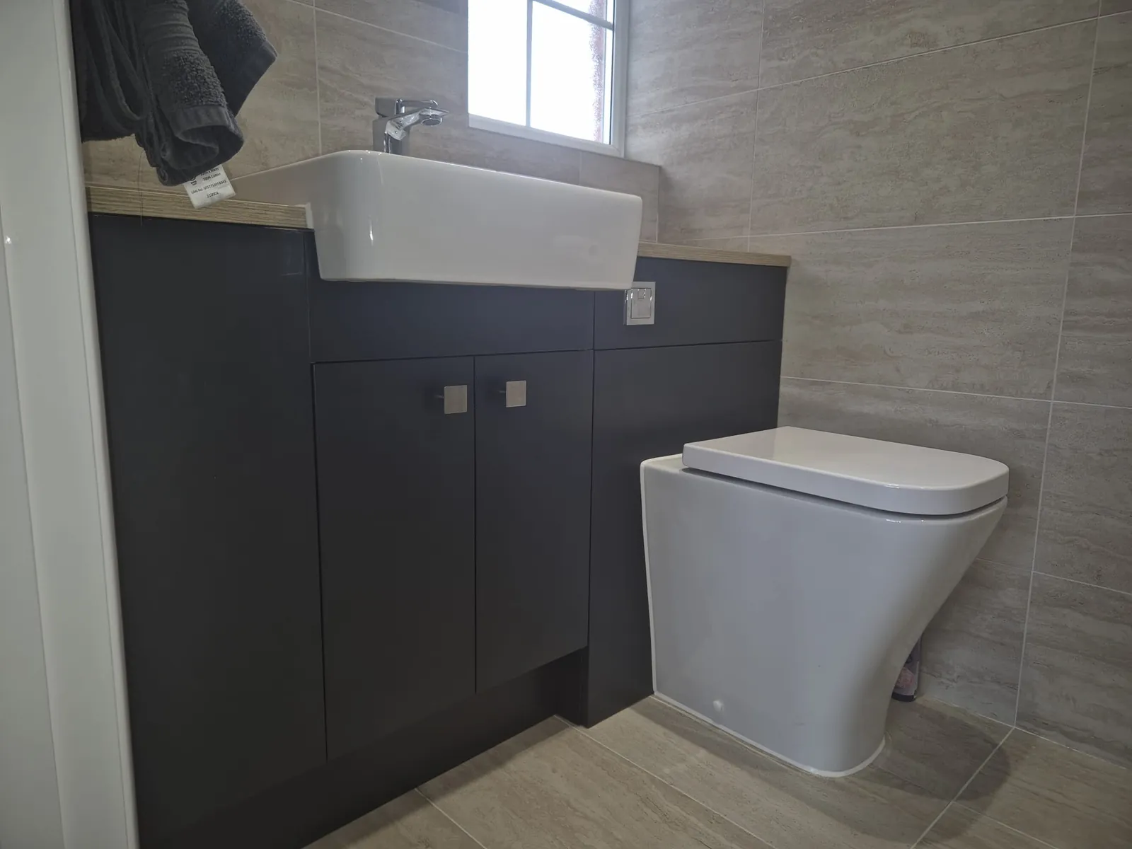 Dark vanity unit with a countertop basin and wall-hung WC in a grey tiled bathroom by GJS