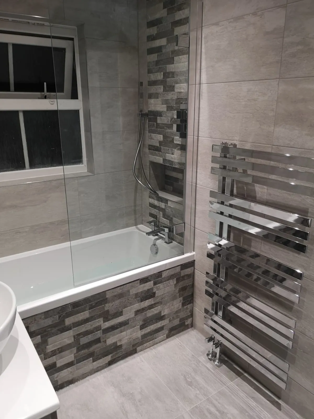 Panelled bath with a chrome heated towel rail and mosaic feature tiling in a grey bathroom by GJS