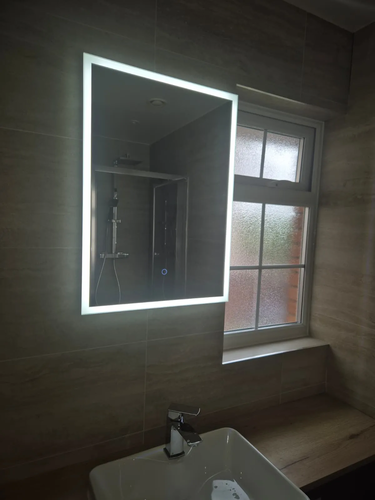Illuminated LED bathroom mirror above a basin in a grey tiled bathroom by GJS