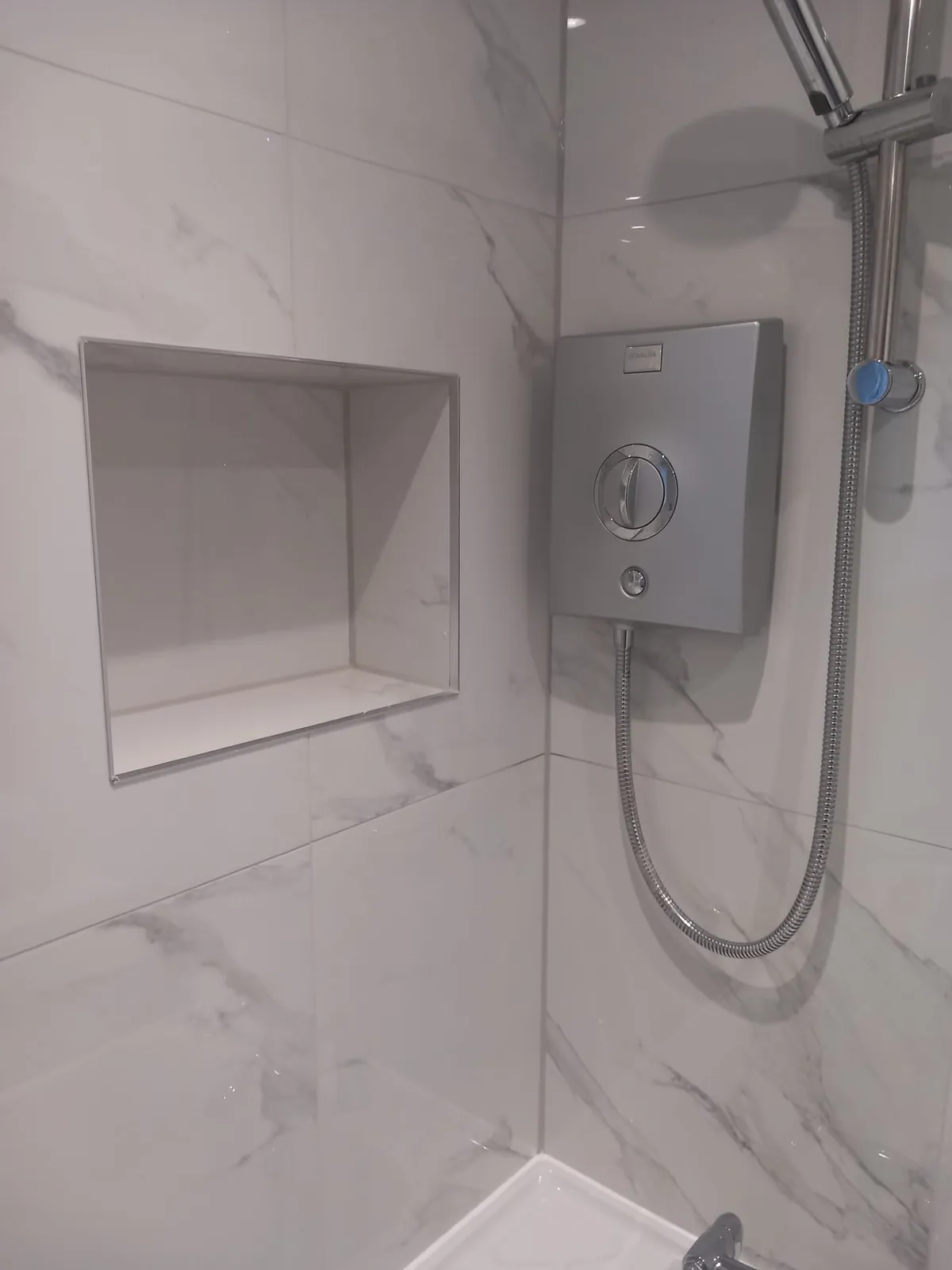 Electric shower and a recessed storage niche in a white marble-effect tiled bathroom by GJS